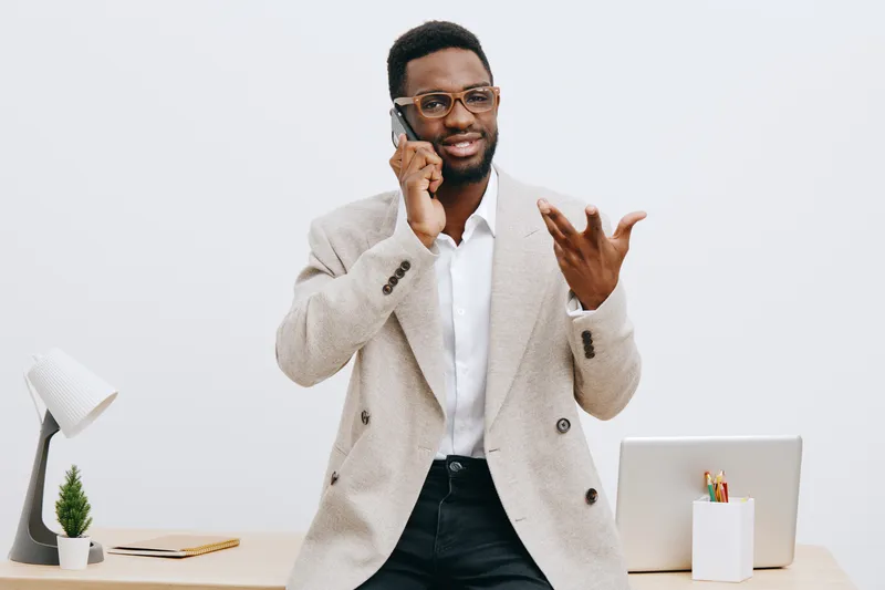 Confident young African-American man in beige blazer talking on smartphone, gesturing with hand, smiling while standing at modern office desk on plain white background, lifestyle and business concept