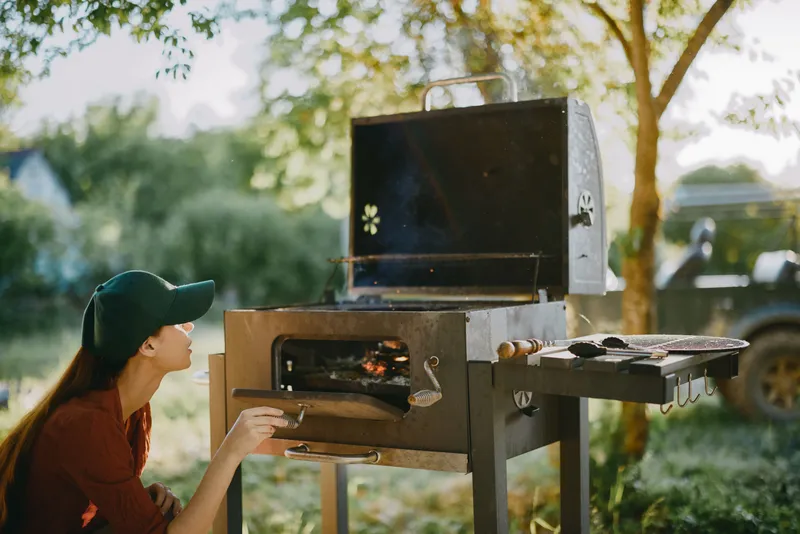 Young woman grilling outdoors in a green environment, wearing a cap and enjoying the barbecue experience, showcasing relaxation and summer vibes