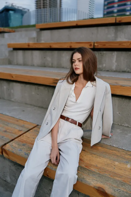 Confident businesswoman seated on wooden steps, wearing elegant cream outfit, exuding professionalism and poise in an urban setting.
