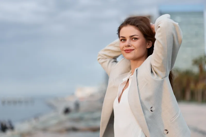 confident woman in light blazer posing outdoors with relaxed smile and natural makeup against soft urban background