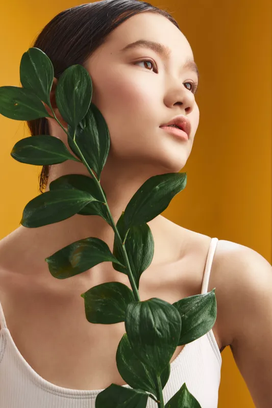 Asian woman gazes softly, holding a leafy plant close in a bright studio, warm backdrop, serene expression, and natural lighting creating a calm, modern aesthetic.