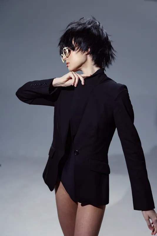 Stylish woman in black blazer with sunglasses posing against gray background expressing confidence and modern fashion.