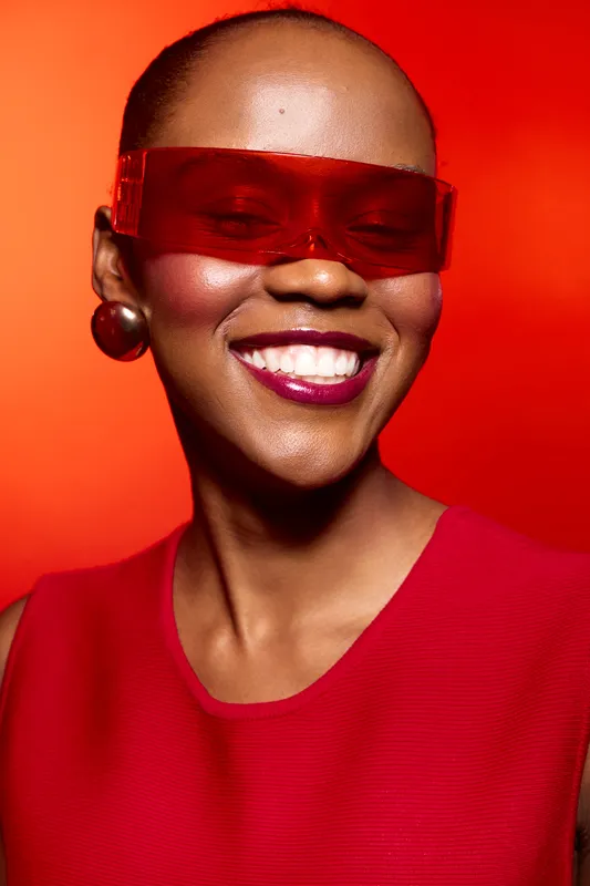 Smiling black woman with dark skin wears red dress and large earrings with red futuristic glasses on vibrant red background. Concept of fashion, beauty, and confident African model.