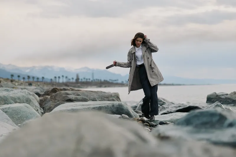 Young woman in long coat walking on rocks by the sea on a cloudy day. Casual outfit and natural landscape with calm water and distant mountains in background.