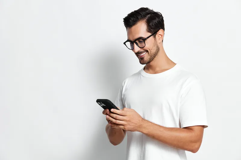 Smiling young man wearing glasses and a white t-shirt, looking at his smartphone with a cheerful expression, isolated on plain light background. Lifestyle concept