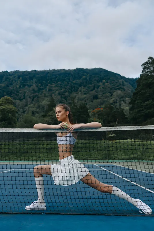 Athletic young woman in stylish tennis outfit poses on outdoor court, showcasing strength and determination with a scenic mountain backdrop