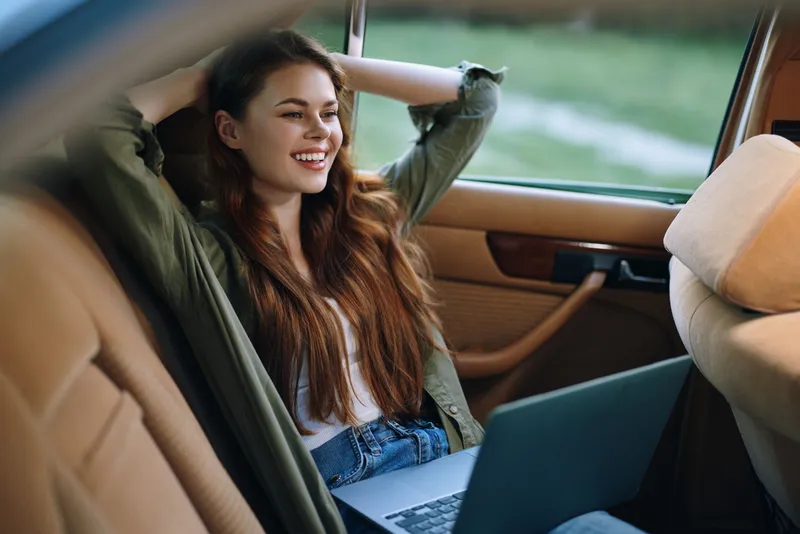 Smiling young woman working on a laptop in a car, enjoying a relaxed atmosphere and sunny day, showcasing happiness, creativity, and modern lifestyle.