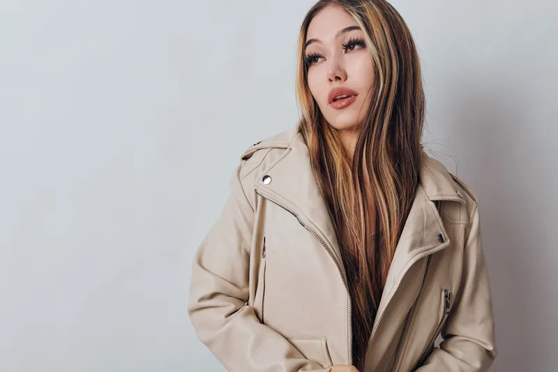 Young woman with long hair wearing beige jacket poses against white wall looking sideways with thoughtful expression in casual fashion style studio portrait.