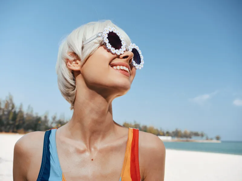 Bright portrait of a blonde woman on a sunny beach, wearing ornate white framed sunglasses and a colorful swimsuit, smiling toward the sky.
