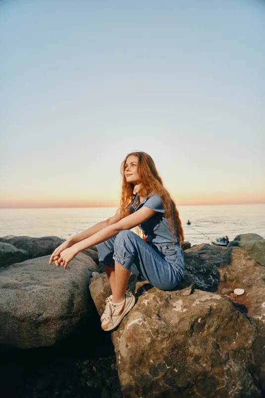 young woman sitting on rocks by the sea during sunset wearing casual denim jumpsuit and sneakers in relaxed outdoor setting with calm ocean horizon