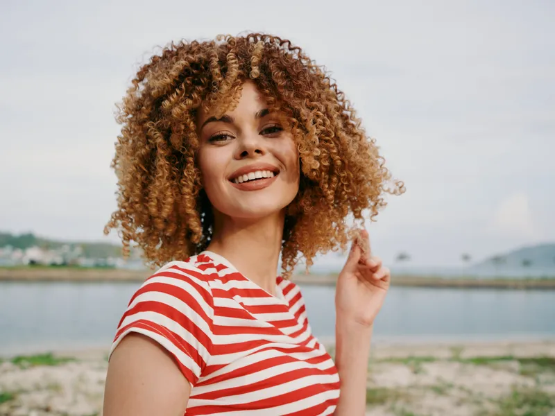 Sunlit portrait of a smiling woman with voluminous curly hair at the seaside, wearing a red and white striped shirt, vibrant and engaging.