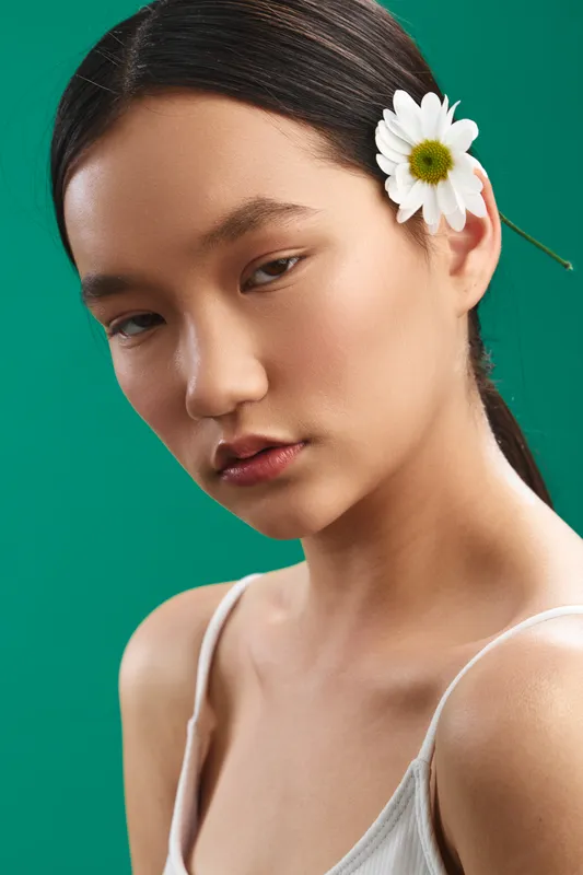 Asian woman wearing a delicate flower in her hair, posed against a green studio backdrop, conveying calm elegance and contemporary beauty in a minimalist portrait.