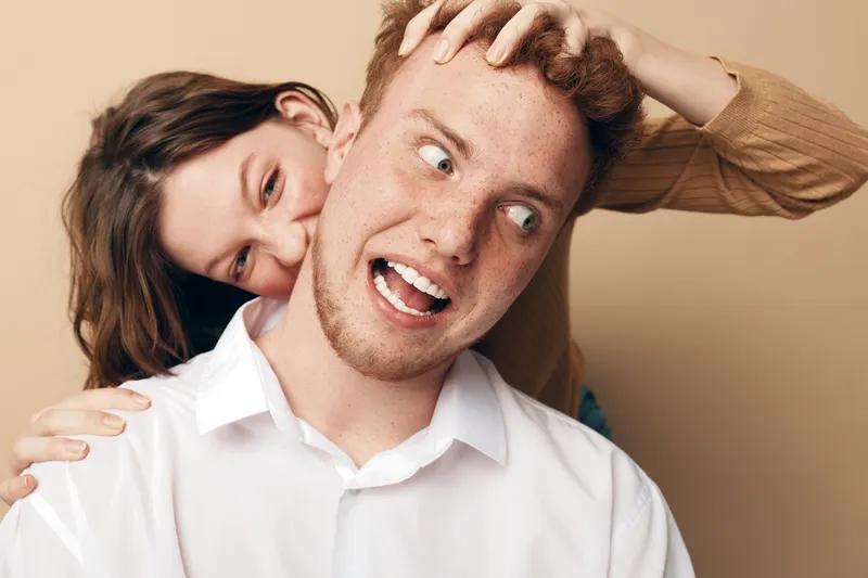 Playful young woman with brown hair teasing redhead man in white shirt on beige background. Concept of fun, friendship, and joyful interaction between friends or couple.
