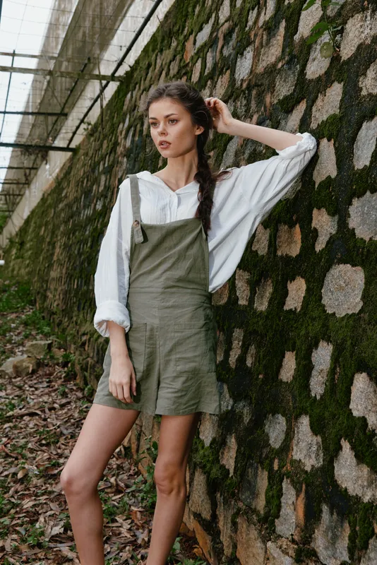Young female model in casual summer outfit with green overalls and white blouse, expressing a relaxed emotion against a natural stone wall backdrop