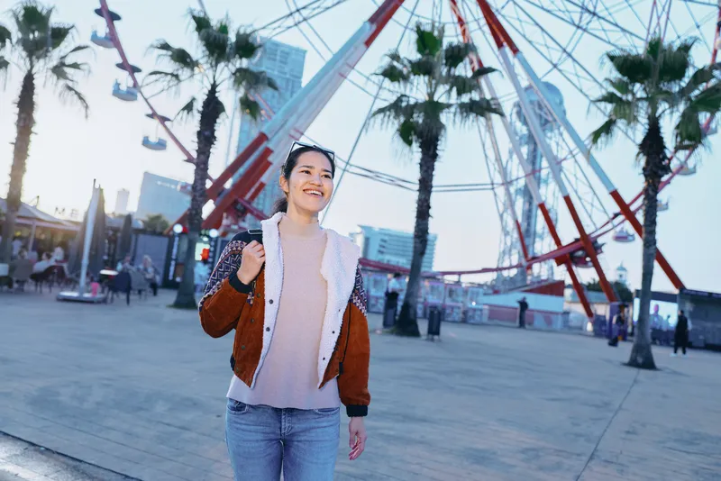 Woman in casual jacket at outdoor plaza near ferris wheel, smiling, capturing urban lifestyle, travel and leisure moment under sunny weather with palm trees and modern architecture