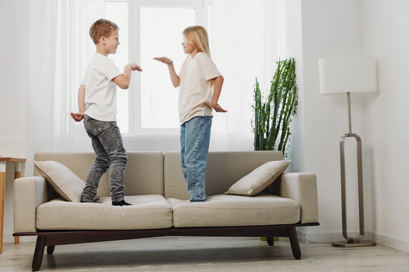 Two kids playfully standing on a beige sofa in a bright living room with casual clothes, interacting with each other and enjoying a fun moment at home with natural light.
