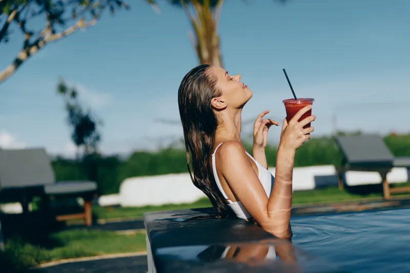 Relaxed woman lounges poolside on a sunny day, sipping a red drink with straw, enjoying leisure moments by the water, capturing a lifestyle scene of summer calm and refreshment.