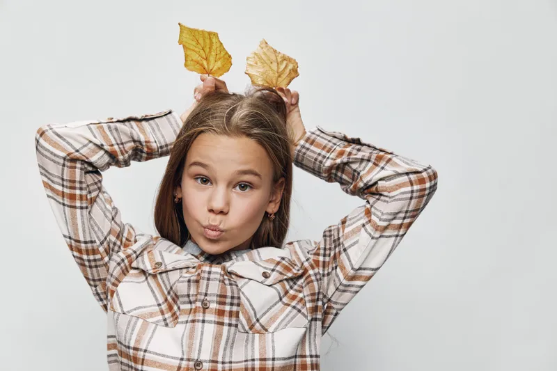 Girl with brown hair holding yellow autumn leaves over head, making playful face in plaid shirt. Child posing with seasonal nature elements on white background.