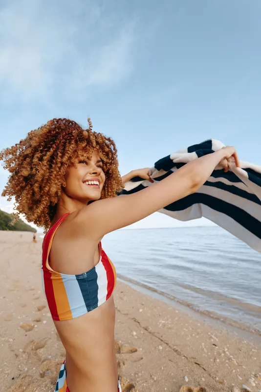 Young woman with curly hair enjoys a sunny day on the beach while holding a striped towel above her head and smiling with a joyful expression.