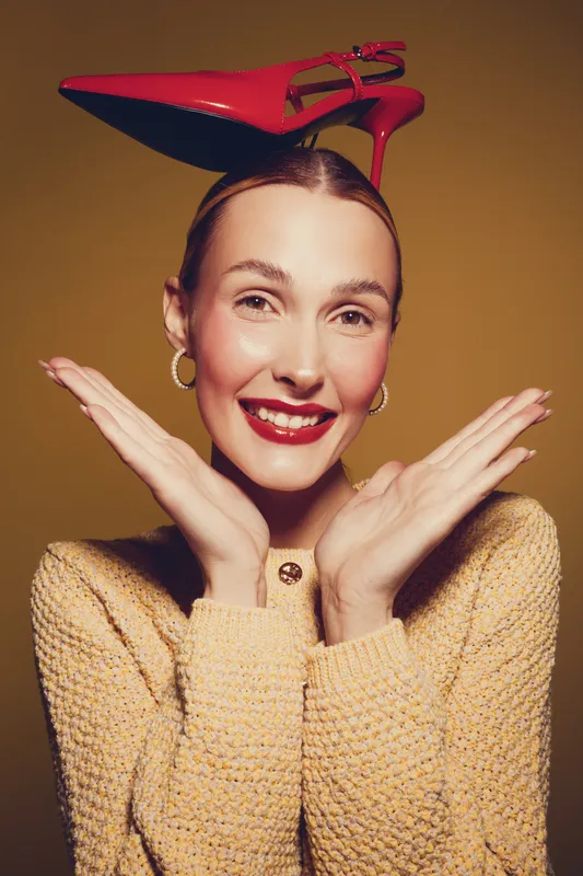 Smiling woman with red high heel shoe on head poses with hands near face. Fashion concept with playful mood and bright lipstick on warm brown background.