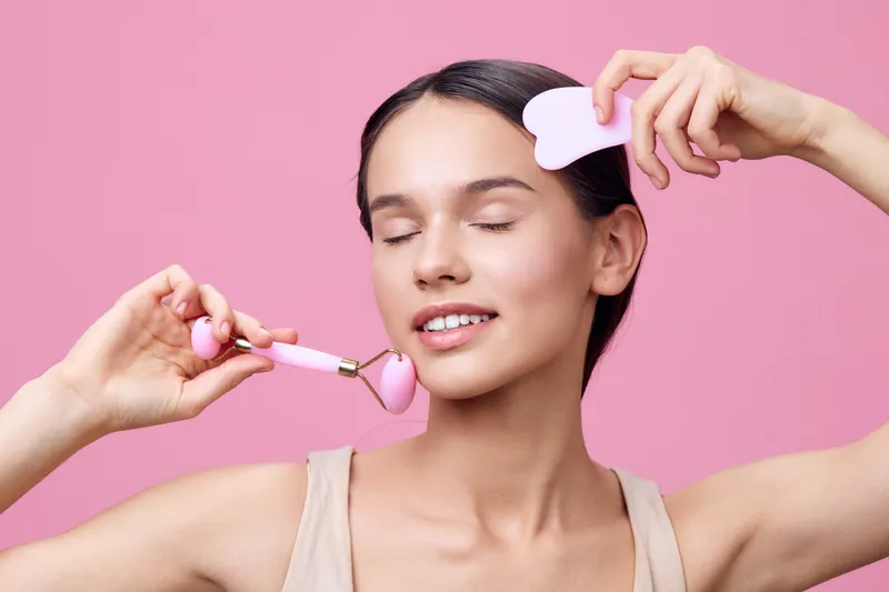Young woman using a jade roller for facial massage, showcasing self care and beauty ritual on a soft pink background