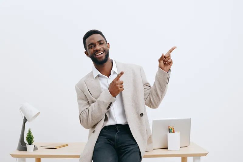 Smiling happy young African-American man wearing beige blazer and white shirt, pointing up with finger, looking at camera, standing in modern office with minimal decor and white background. People lif