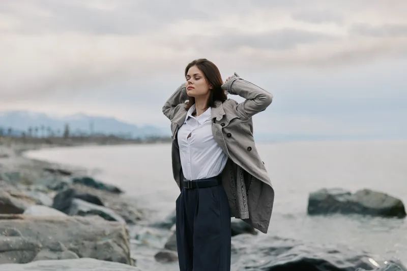 Young woman in a stylish coat stands on rocky shore looking away thoughtfully. Overcast sky and calm sea create a serene natural outdoor atmosphere. Concept of fashion and contemplation.