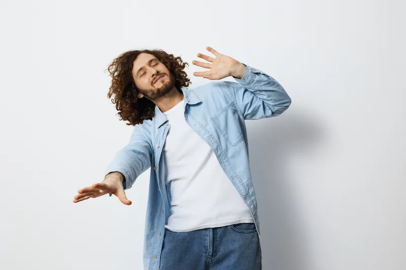 Young man dancing joyfully with curly hair in casual clothes, expressing happiness against a light background