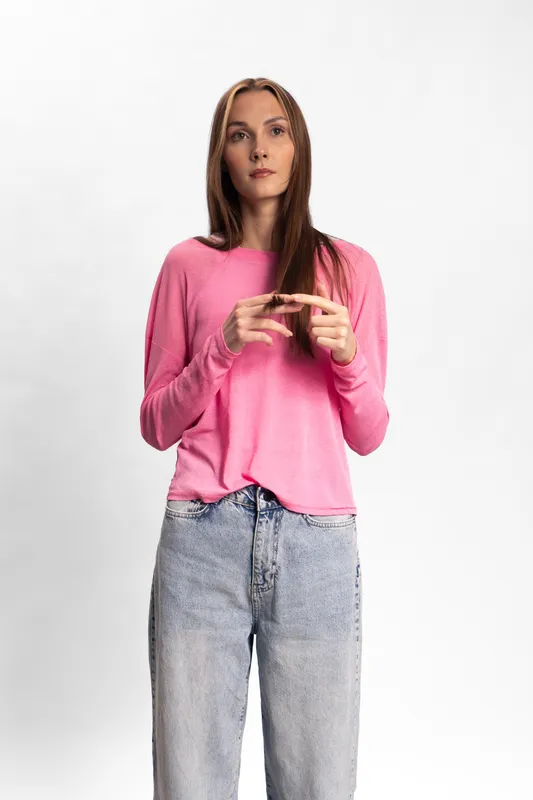 A young woman with long brown hair wearing a pink sweater and light blue jeans stands against a white background. She has a neutral expression and holds her hands together. Casual fashion concept.