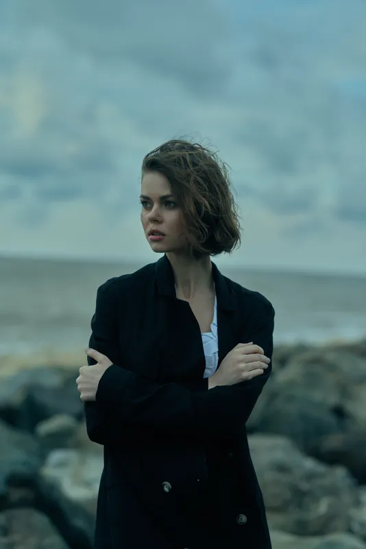 Woman in a dark coat stands with crossed arms beside a rocky coastline, moody sea backdrop, conveying introspection, fashion, and strength