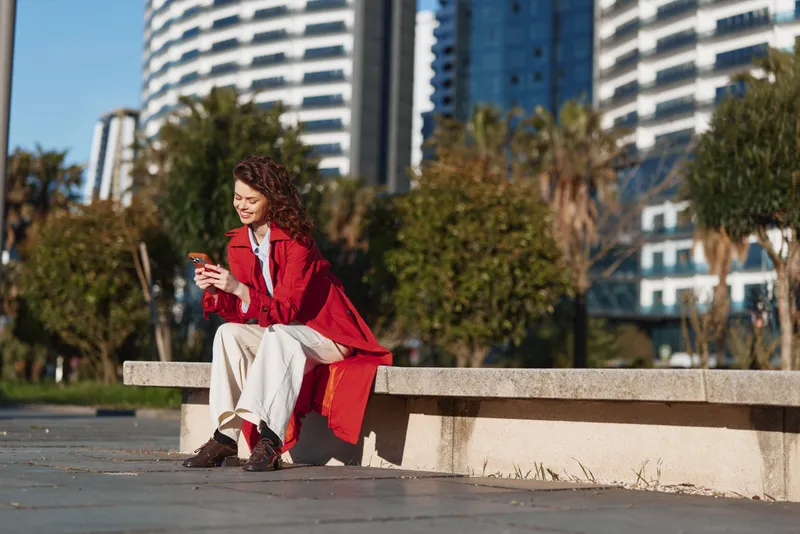 Young woman in red coat sits on a concrete bench in a sunny urban park, scrolling on her phone with a relaxed pose, a candid moment
