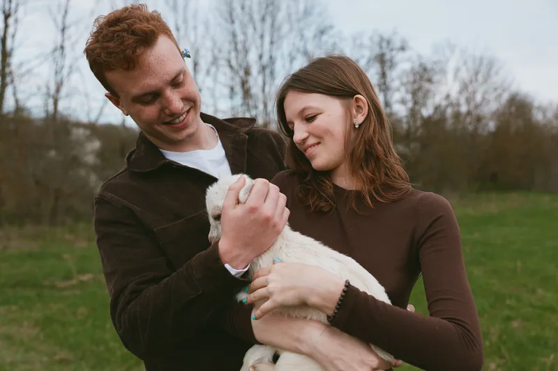 Cheerful couple outdoors, gently cradling a small goat kid, warm sunlight, casual sweaters, relaxed mood, rustic trail backdrop, candid moment of tenderness, fresh air, natural colors, joyful