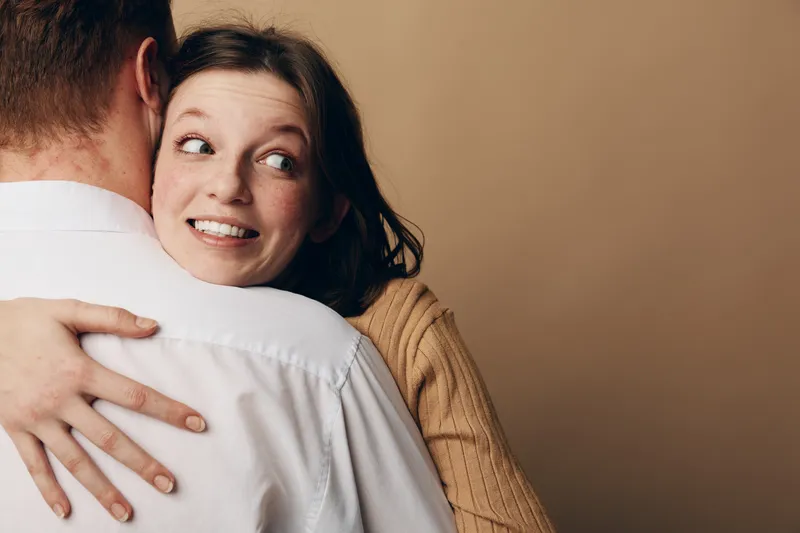 Smiling woman with freckles hugs man wearing white shirt, looking sideways with happy expression. Intimate moment of affection and connection between couple in warm indoor setting.