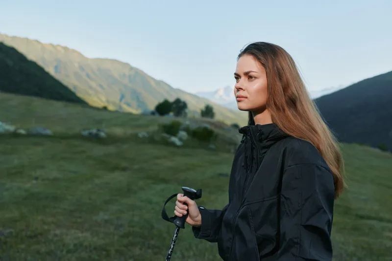 A thoughtful woman stands in a scenic valley with a trekking pole, wearing a dark jacket. She gazes into the distance, conveying calm focus and a sense of outdoor adventure.