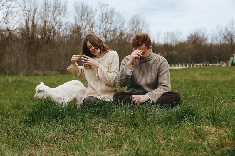 A casual outdoor scene features two adults and a white goat kid on a grassy park, one scrolling a phone while the other rests with a beverage. A candid lifestyle moment centered on animal