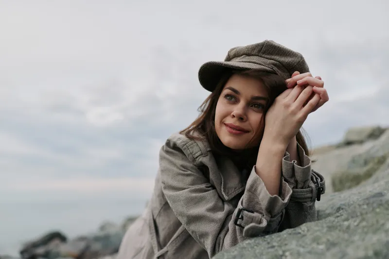 Young woman in casual coat and hat leans on rocky surface outdoors, smiling softly. Overcast sky and sea are visible in the background. Concept of nature, relaxation, and autumn fashion.