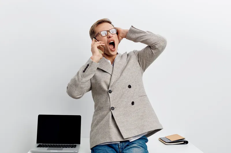Businessman wearing glasses in casual suit sitting at desk with laptop and notepad, shocked or surprised, against plain white background. Office worker emotions concept