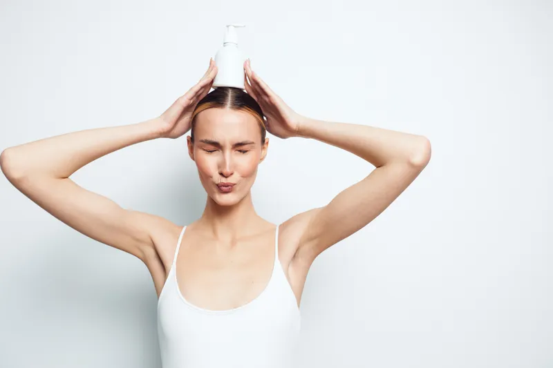 A young woman in a white tank top holds a white lotion bottle on her head with both hands, eyes closed and lips puckered, standing against a plain white background. Concept for skincare and self-care.