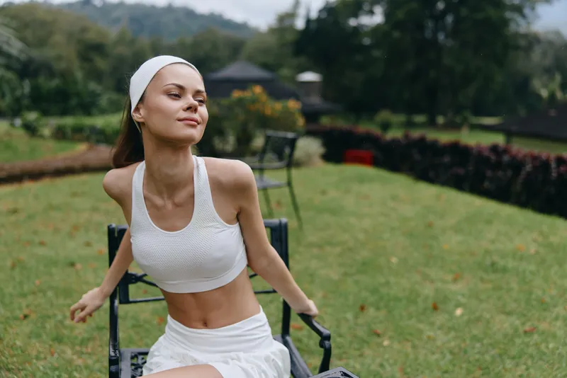 Beautiful young woman practicing yoga outdoors, wearing a white sport outfit, with serene expression in a lush green park setting, promoting wellness and fitness