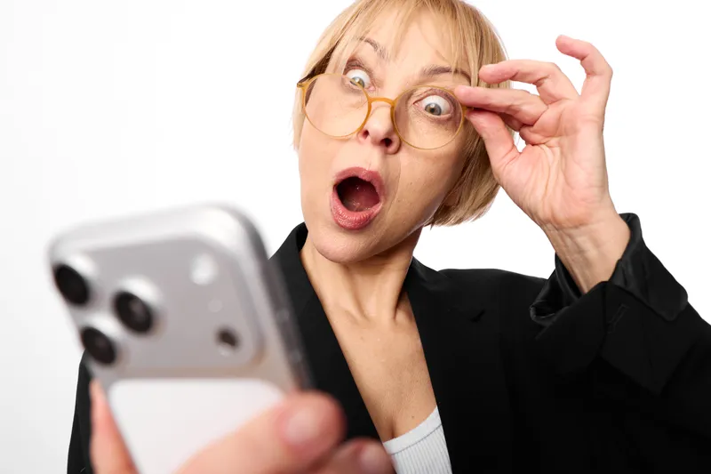 Woman surprised smartphone glasses portrait on white studio background, blonde woman in black blazer holding phone, open mouth and wide eyes, shock communication