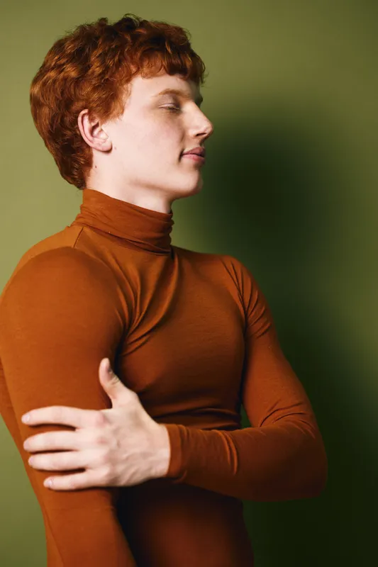Portrait of young man with red hair and freckles wearing brown turtleneck against green background. Side profile, eyes closed and arms crossed in thoughtful pose.