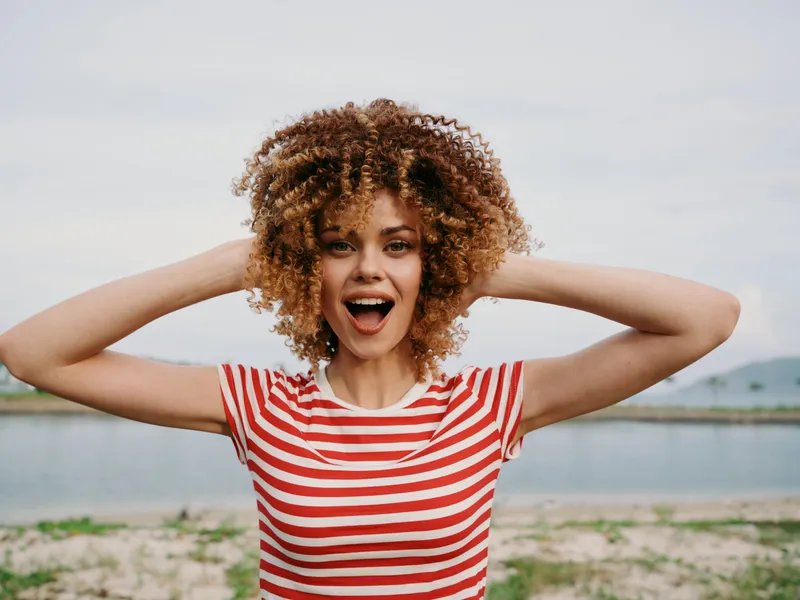 A lively young woman with curly hair wearing a red and white striped shirt stands on a sunny beach, expressing excitement with hands near her head and a bright smile.
