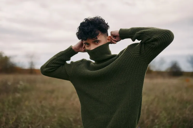 In a windswept field a young man pulls a green knit sweater over his face, a candid portrait emphasizing natural expression, texture and authenticity and credibility in an intimate moment.