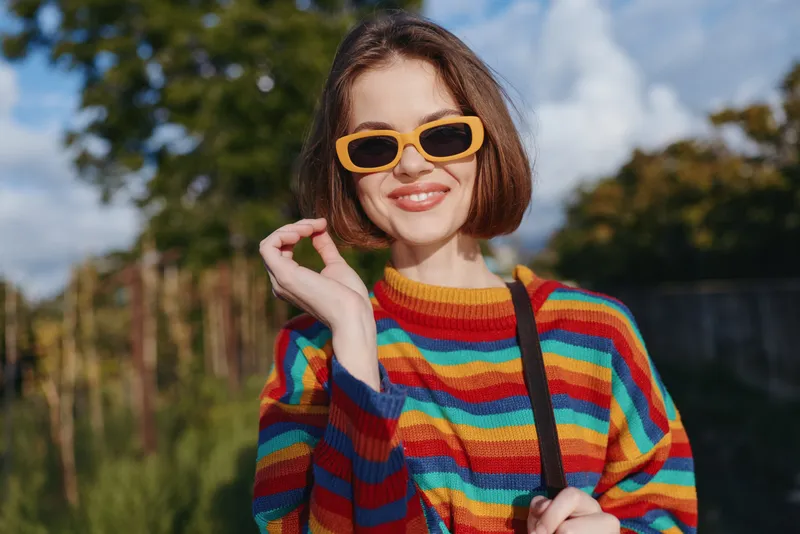 Young woman in her 20s wearing a colorful striped sweater and yellow sunglasses, smiling portrait outdoors with short bob haircut, casual bag strap and sunny lifestyle vibe.
