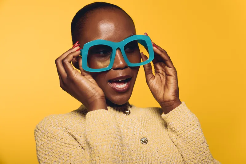 Confident black woman with dark skin wearing oversized blue glasses and beige sweater poses against yellow background. Concept for fashion, style, and beauty with vibrant colors.