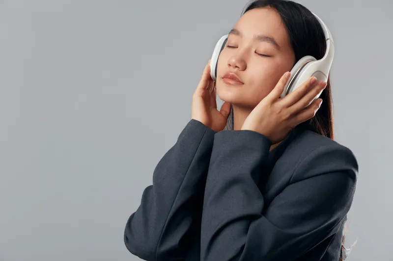 Young woman with long hair wearing headphones and dark jacket, eyes closed, enjoying music or relaxing in studio with neutral gray background and calm expression.