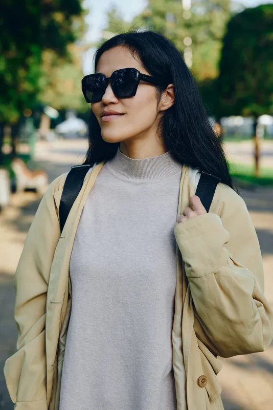 Woman sunglasses coat outdoors park fashion casual sweater, jacket garment apparel streetwear daylight cityscape scenery lifestyle accessory backpack pose confident stylish relaxed urban midday
