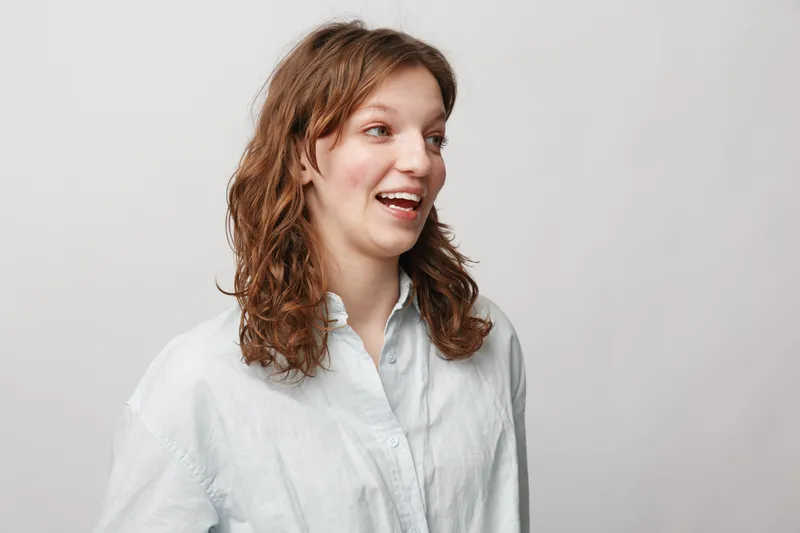 Young woman with curly brown hair wearing a white shirt smiles and looks to the side against a plain light background. Casual portrait with natural expression and soft lighting.