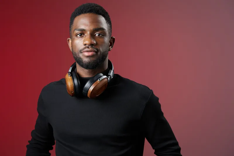 Confident Black man with headphones, wearing a black sweater, smiling against a red background, representing music enjoyment and modern lifestyle