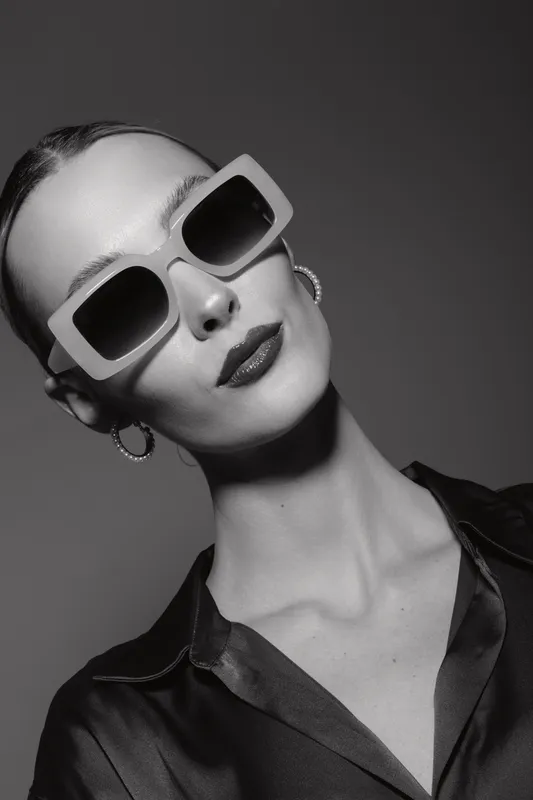 Portrait of a fair skinned caucasian woman wearing large sunglasses and hoop earrings. She poses confidently with slicked back hair and a dark satin shirt in studio lighting.